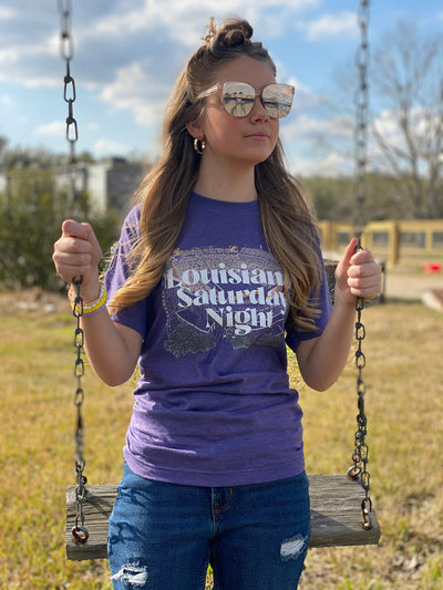 Brunette model wearing sunglasses sitting on a swing. Shirt is a heather purple tee with the LSU stadium in yellow and the words Louisiana Saturday Night in White font over the stadium