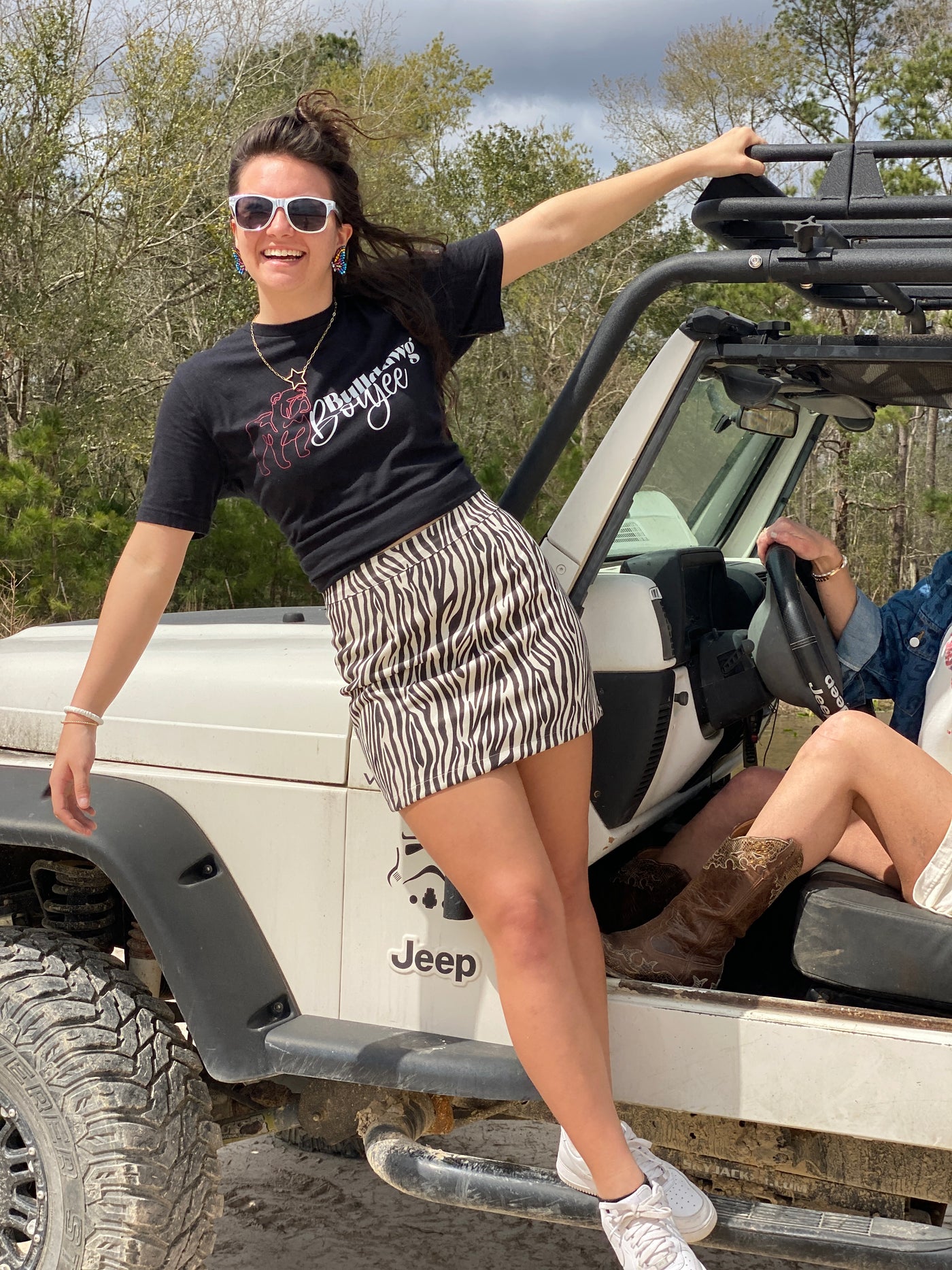 Brunette model hanging off a white jeep in a zebra print skirt and a black shirt that has a line drawing of a bulldog in red and the words Bulldog Boujess in white next to the bulldog and the word boujee is in cursive