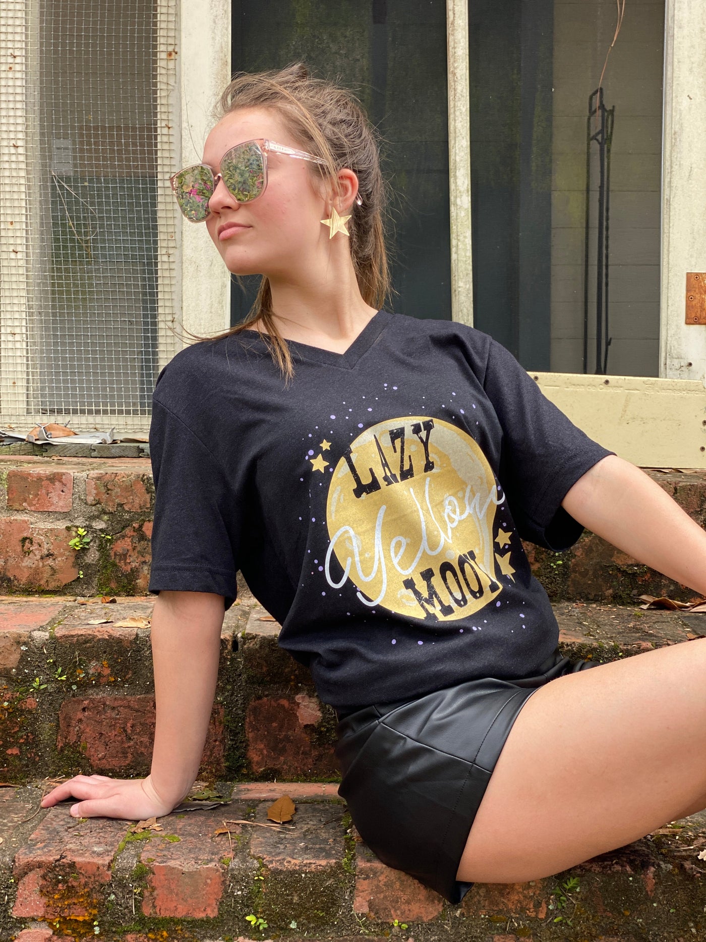 Heather black V neck shirt with a the graphic of a yellow moon surrounded by yellow stars and white dots. The words Lazy Yellow Moon are across the moon Lazy and Moon are in a western vintage font and yellow is in a white script font 