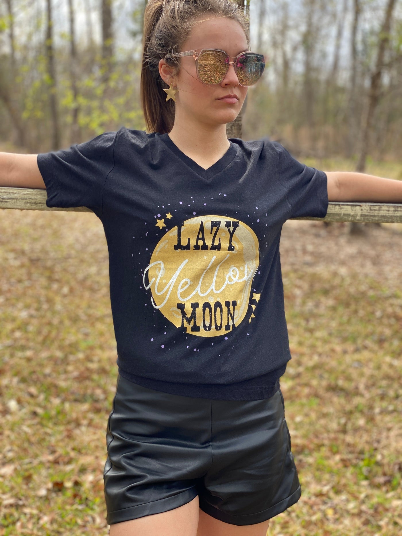 Heather black V neck shirt with a the graphic of a yellow moon surrounded by yellow stars and white dots. The words Lazy Yellow Moon are across the moon Lazy and Moon are in a western vintage font and yellow is in a white script font