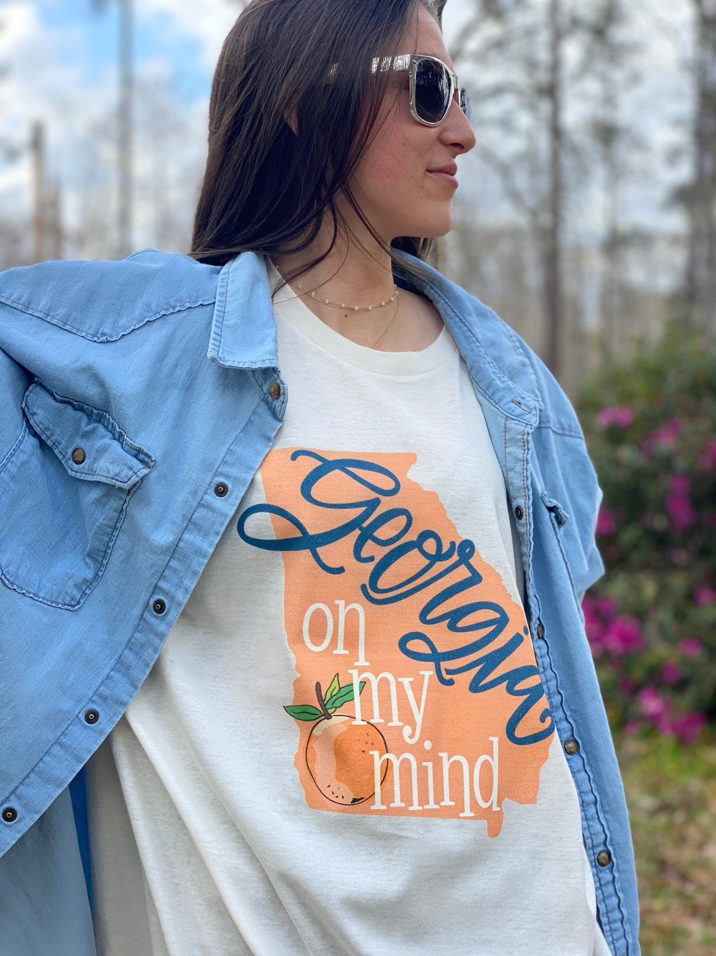 Brunette model wearing sunglasses and a cream graphic tee under a denim shirt. The graphic on the tee is the state of Georgia in peach with the words Georgia on my mind and a peach on the Georgia. The Georgia is a blue cursive font and on my mind is in white.
