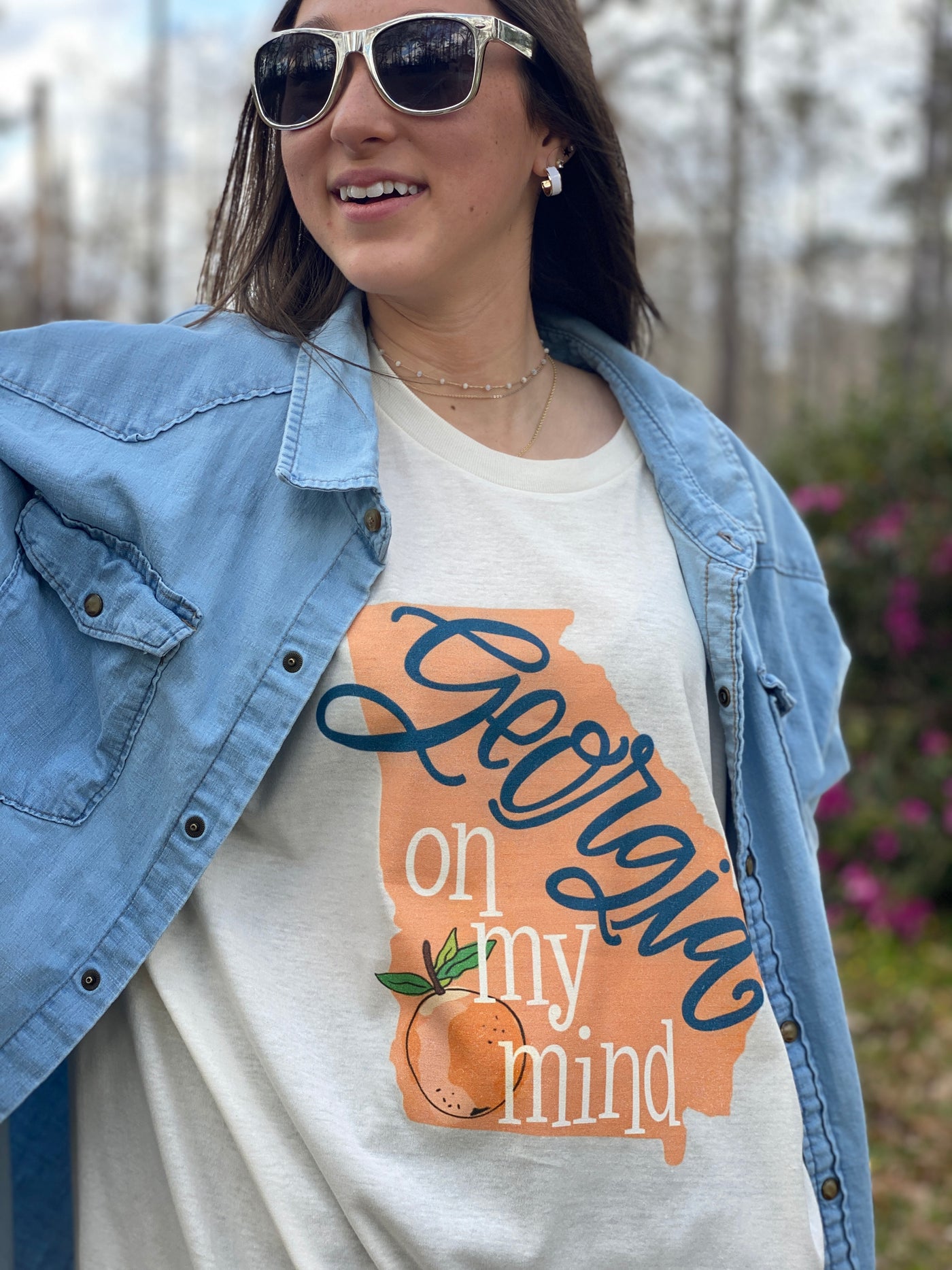 Brunette model wearing sunglasses and a cream graphic tee under a denim shirt. The graphic on the tee is the state of Georgia in peach with the words Georgia on my mind and a peach on the Georgia. The Georgia is a blue cursive font and on my mind is in white.