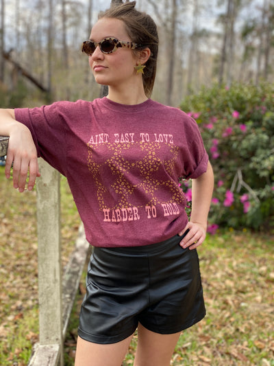  Red tee with graphic. Graphic is Pink weathered letters at the top read " Aint Easy To Love" underneath the text there are 9 different styles of cowboy hats outlined in brown with a tan leopard print in between the cowboy hats, text below the hats reads " Harder To Hold"