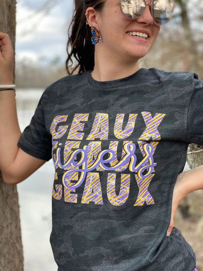Brunette model wearing large sunglasses standing in between two trees in a graphic shirt. Shirt is a dark camo color with a Graphic that says "Geaux Geaux Geaux" that is stacked and has a graffiti print in yellow purple and white and the word tigers in a light purple cursive font over the top of the stacked Geauxs