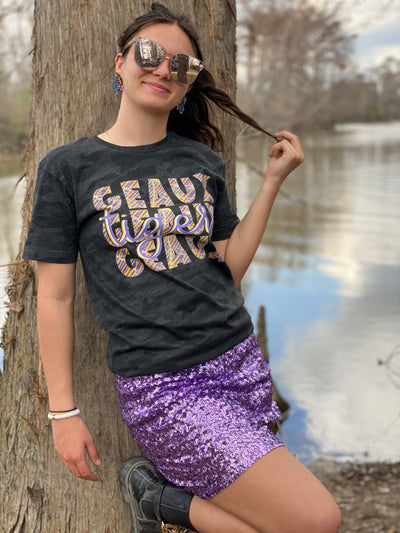 Brunette model wearing large sunglasses standing in between two trees in a graphic shirt and a purple sequin skirt. Shirt is a dark camo color with a Graphic that says "Geaux Geaux Geaux" that is stacked and has a graffiti print in yellow purple and white and the word tigers in a light purple cursive font over the top of the stacked Geauxs.