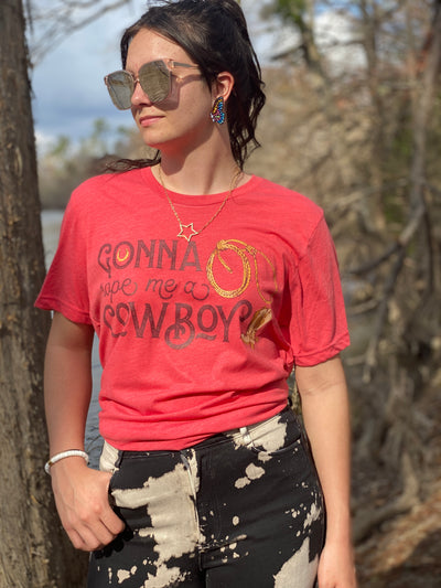 a red shirt with the words "Gonna Rope me a Cowboy" In a western font. In the O of gonna there is a horseshoe and to the right there is a rope and a cowboy boot.