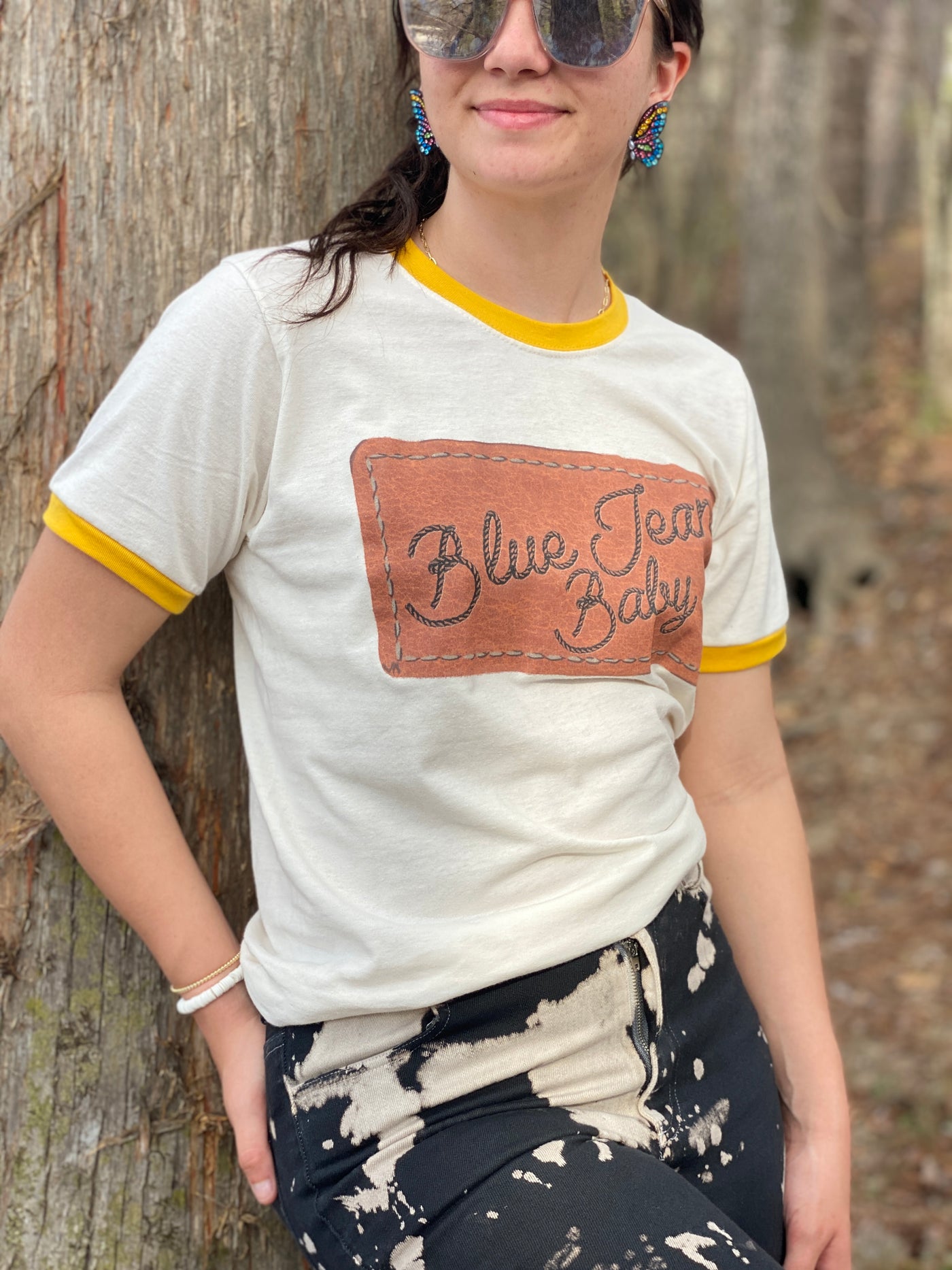 Brunette Model wearing a yellow and cream ringer tee and black and white bleach splatter pants. Tee has a graphic that looks like a leather patch sewn onto the shirt and the words Blue Jean Baby in rope font on the patch.