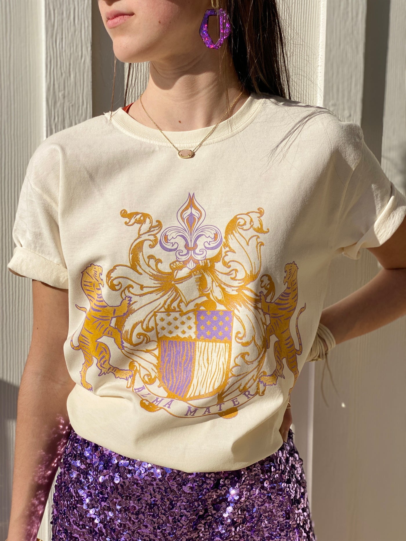 Cream tee. Alma Mater Classic family crest style hand drawn design.  Crest has a tiger on each side with a banner below that reads Alma Mater with a fleur de Lis with filigree on top. Design is in LSU Purple and Gold