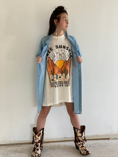 Tan Shirt with a desert scene. There is a sun setting between two rock formations with cactus in the foreground above the graphic says free sunsets and bellow it says no filter needed shine on. Worn oversized as a dress under a denim shirt with fringe, and fringe cowprint cowboy boots.