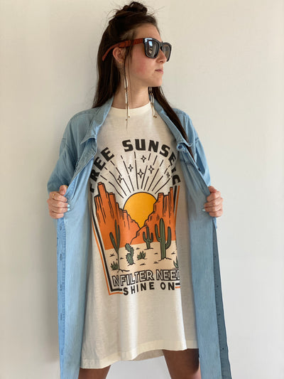 Tan Shirt with a desert scene. There is a sun setting between two rock formations with cactus in the foreground above the graphic says free sunsets and bellow it says no filter needed shine on. Worn oversized as a dress under a denim shirt with fringe.
