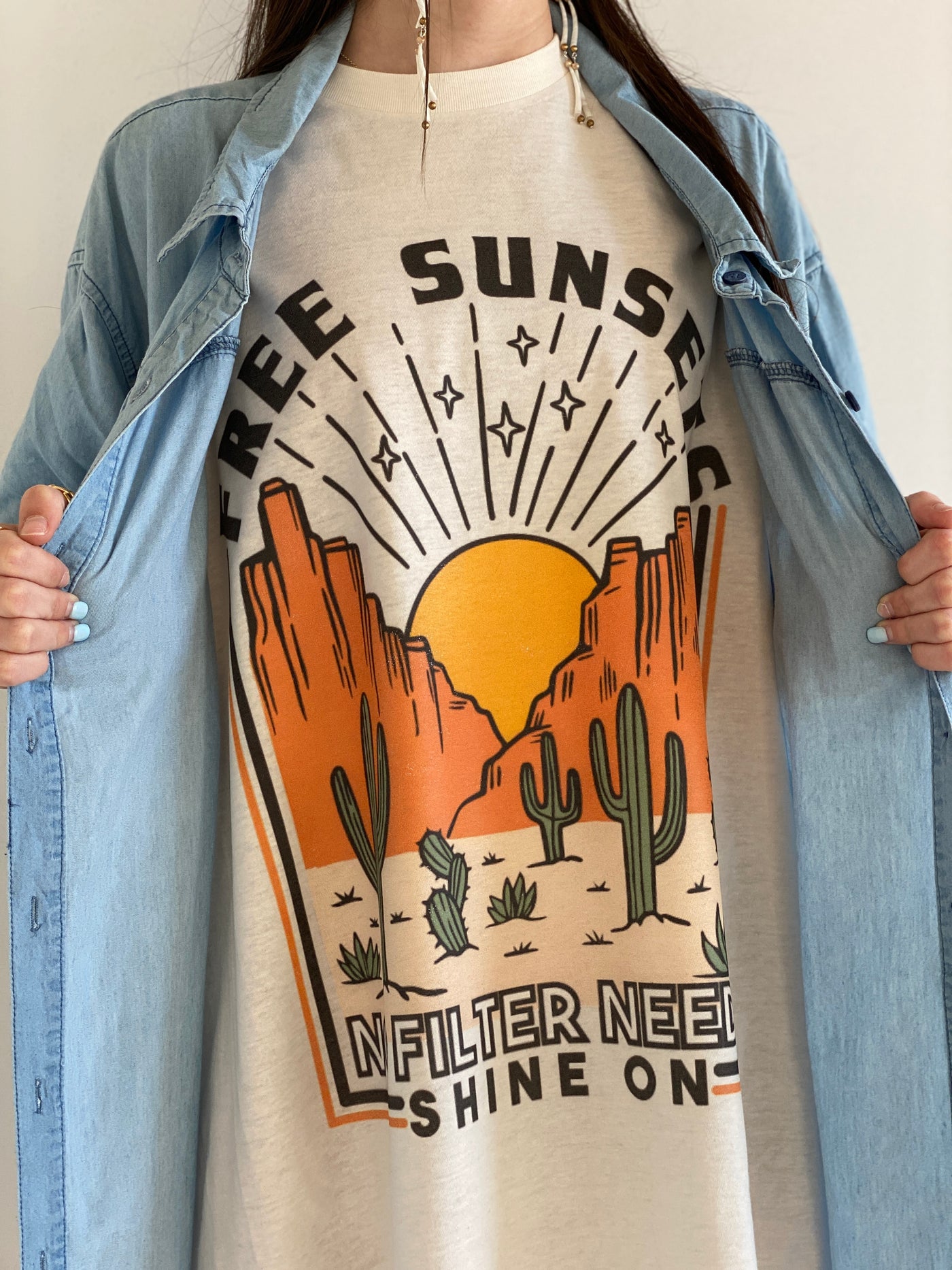 Tan Shirt with a desert scene. There is a sun setting between two rock formations with cactus in the foreground above the graphic says free sunsets and bellow it says no filter needed shine on. Worn oversized as a dress under a denim shirt