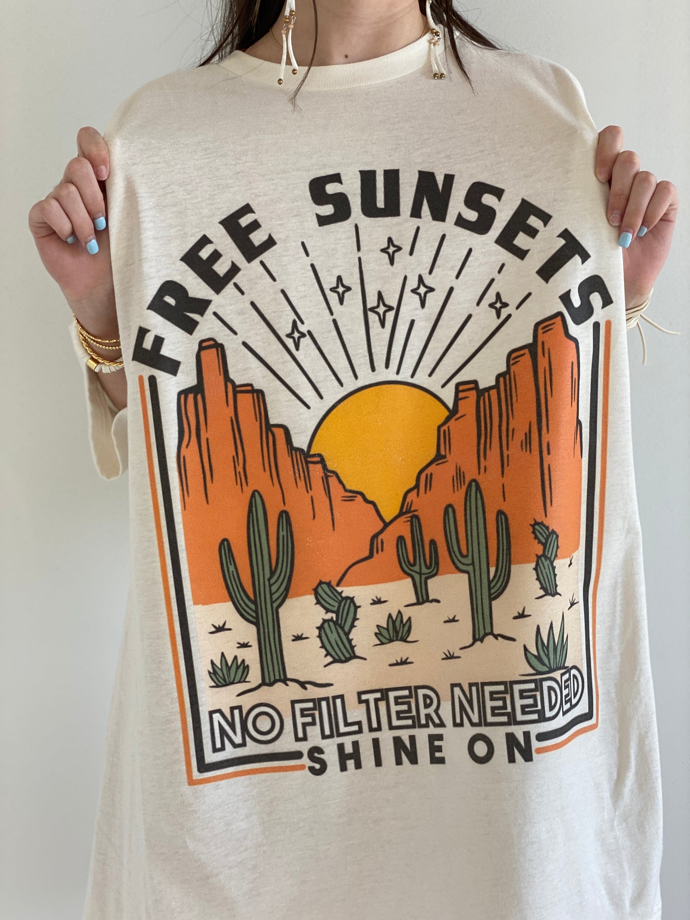 Tan Shirt with a desert scene. There is a sun setting between two rock formations with cactus in the foreground above the graphic says free sunsets and bellow it says no filter needed shine on. Worn oversized as a dress.