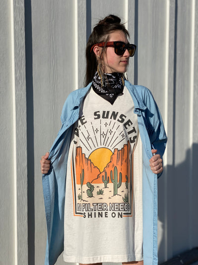 Tan Shirt with a desert scene. There is a sun setting between two rock formations with cactus in the foreground above the graphic says free sunsets and bellow it says no filter needed shine on. Worn oversized as a dress under a denim shirt with fringe.