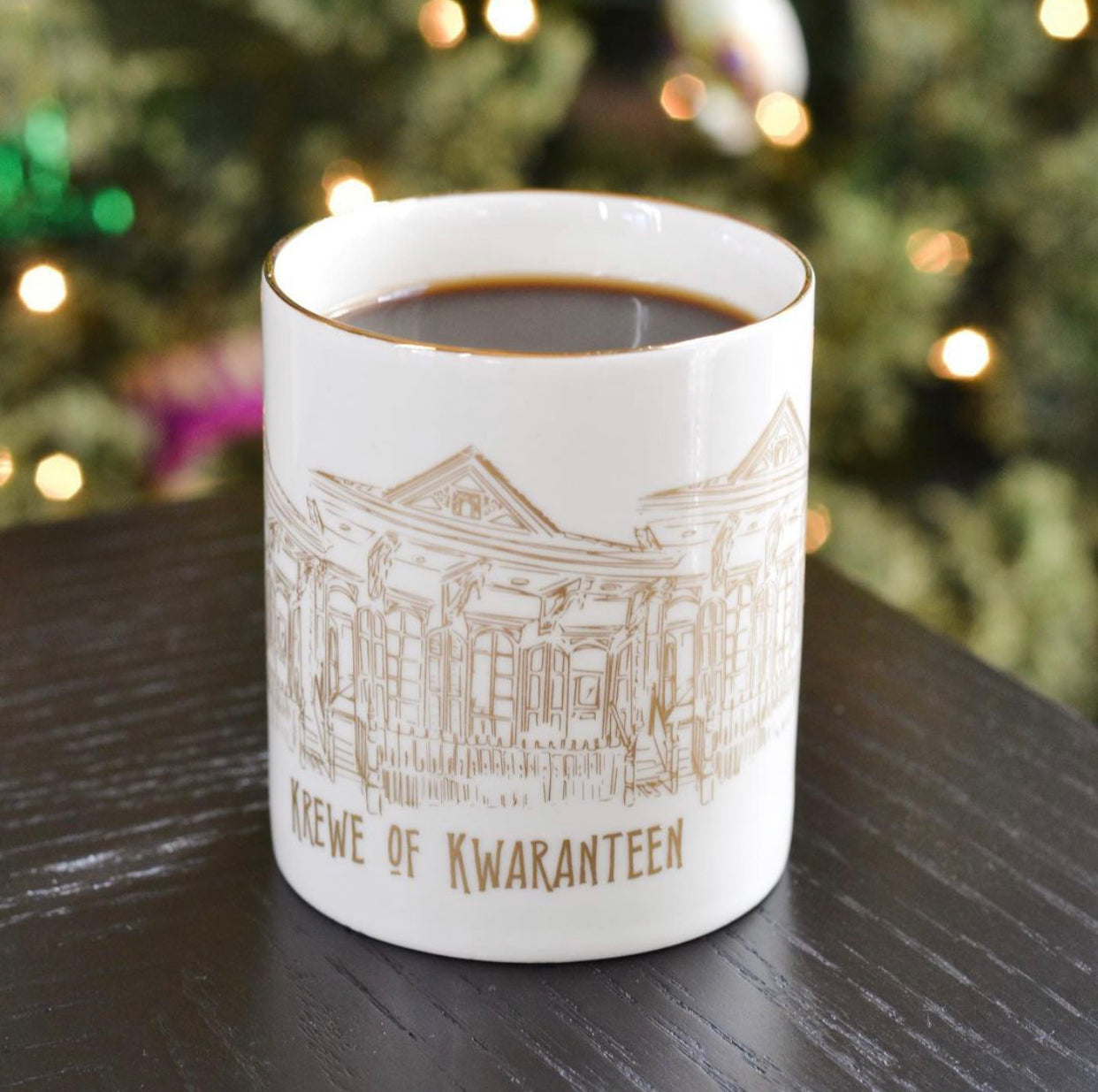 A white coffee cup with a gold rim and the graphic of historic New Orleans houses and the words Krewe of Kwaranteen underneath in gold
