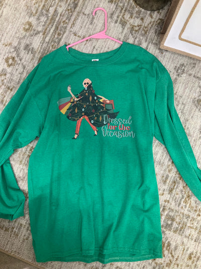 Green long sleeve tee with graphic. graphic is of a woman wearing a Christmas tree with lights and ornaments, SHe has on red pants and black high heels and round sunglasses. She is carrying shopping bags and flashing a credit card. The were Dressed for the Occasion are in the bottom right hand corner of the design the words dressed and occasion are in green cursive font and for the is in a red block font. 