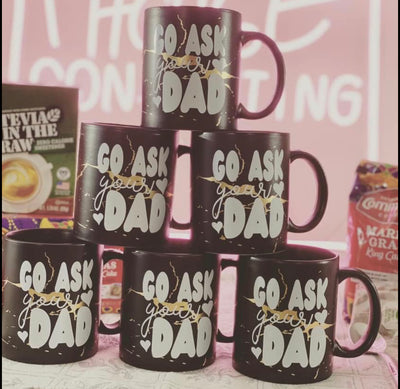 A pyramid of black coffee mugs. A black coffee mug with gold veins. On the coffee mug there are the words "Go Ask" in bubble letter then the word "your" in cursive under that and on the bottom says "dad" in bubble font.