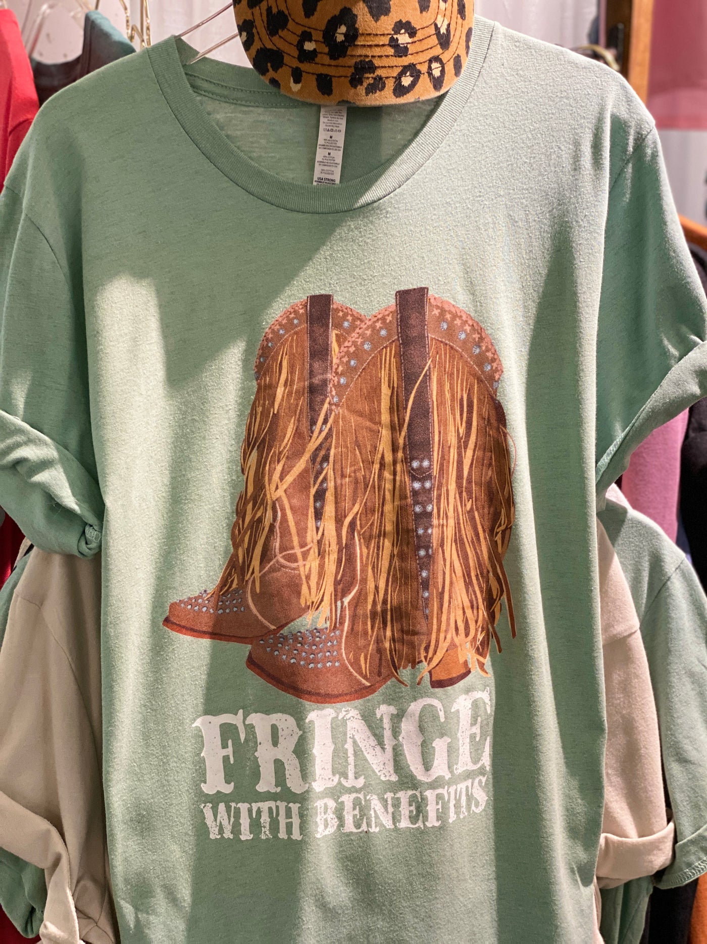 Green tee with graphic. The graphic on the tee is a pair of cowboy boots adorned with rhinestones and fringe and the words fringe with benefits in a western font under the boots.