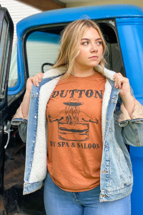 Blonde woman in front of a blue truck wearing blue jeans a denim jacket and a burnt orange shirt with a black graphic on it. The graphic Has the word Dutton in old western letters at the top and Day Spa & Saloon at the bottom, in between the words you can see a womans back with long hair under her cowboy hat sitting in a water trough holding a bottle of whiskey.