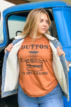 Blonde woman in front of a blue truck wearing blue jeans a denim jacket and a burnt orange shirt with a black graphic on it. The graphic Has the word Dutton in old western letters at the top and Day Spa & Saloon at the bottom, in between the words you can see a womans back with long hair under her cowboy hat sitting in a water trough holding a bottle of whiskey. 