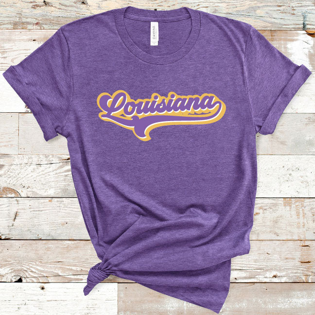 Heather Purple Tee with the word Lousiana in a retro font and swooping tail that goes under the word Louisiana the word has a white outline and a thicker gold outline around thee white.