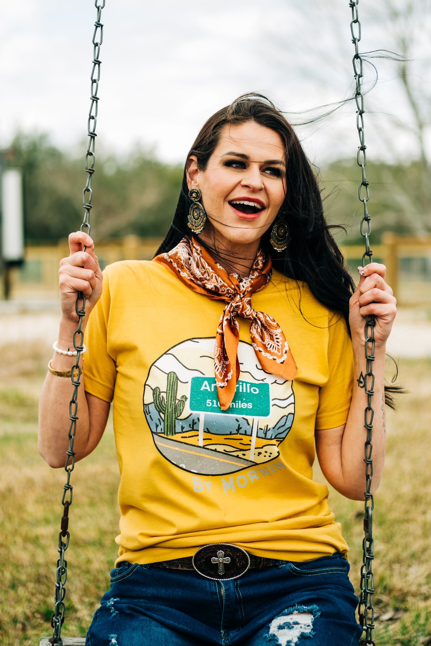Woman Sitting in a swing in a Yellow T shirt that has a circle graphic that contains a desert scene with cactus and a road sign that reads Amarillo 510 Miles. Under the circle has silver letters that read by morning.