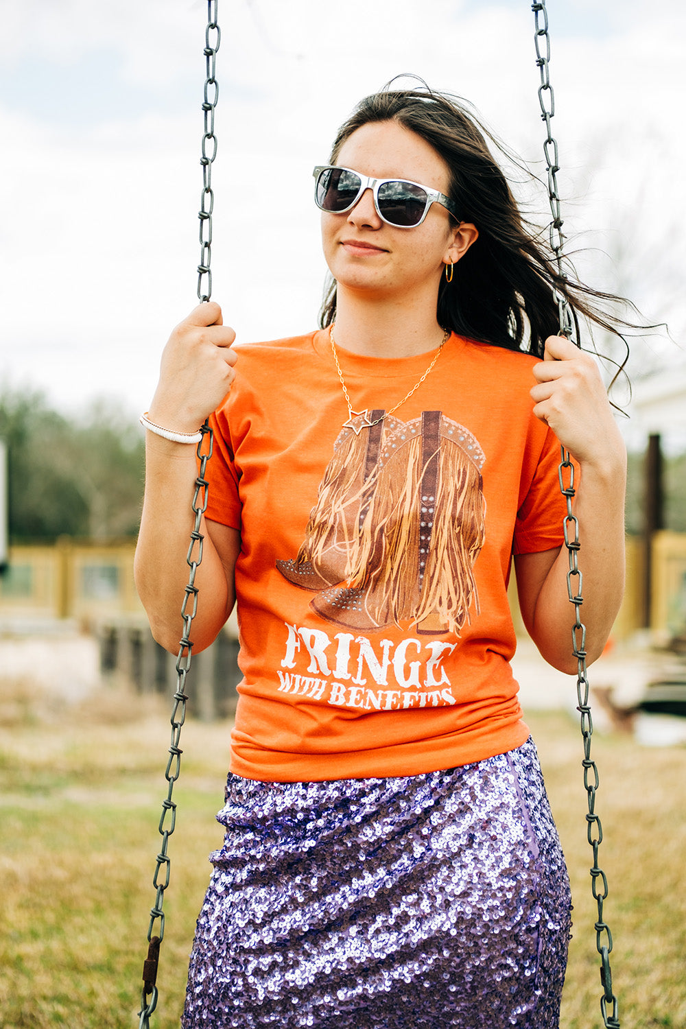 Brunette model in sunglasses and an orange graphic tee and a sequin skirt. The graphic on the tee is a pair of cowboy boots adorned with rhinestones and fringe  and the words fringe with benefits in a western font under the boots.  