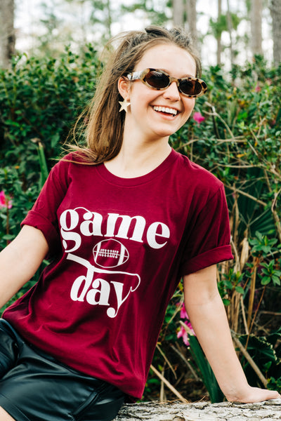 A maroon shirt with the word game stacked on top of a football stacked on top of the word day all in white. 
