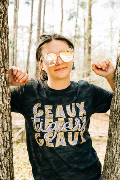 Brunette model wearing large sunglasses standing in between two trees in a graphic shirt. Shirt is a dark camo color with a Graphic that says "Geaux Geaux Geaux" that is stacked and has a graffiti print in yellow purple and white and the word tigers in a light purple cursive font over the top of the stacked Geauxs