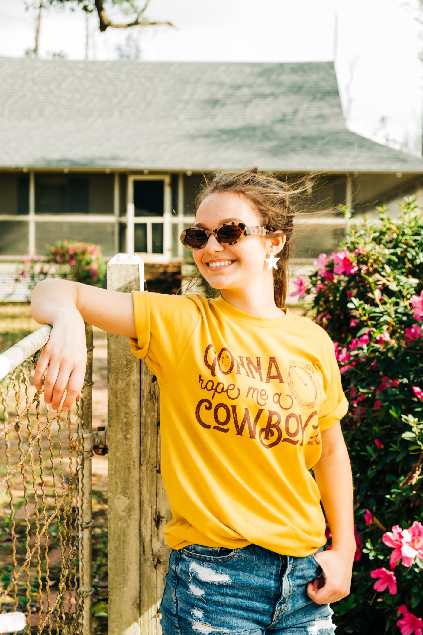 A yellow shirt with the words "Gonna Rope me a Cowboy" In a western font. In the O of gonna there is a horseshoe and to the right there is a rope and a cowboy boot.