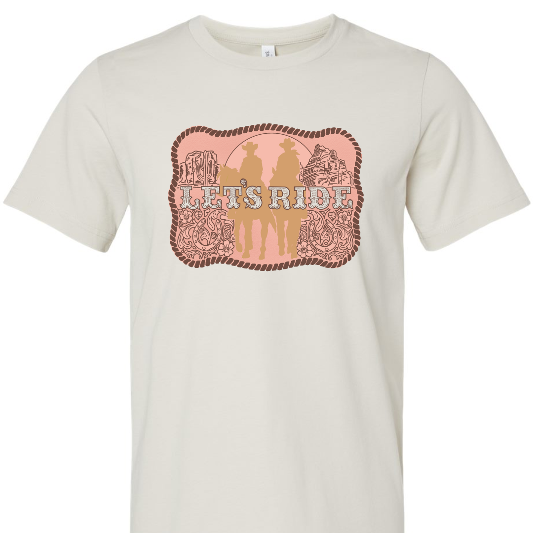 A dust colored tee with a graphic of a belt buckle. Belt buckle is a pink color with a dark brown rope border and brown western filigree on the sides with two cowboys on horses in light tan in the middle of the belt buckle and the words Let's Ride in silver across the width of the belt buckle.