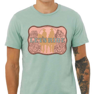 A dusty blue  colored tee with a graphic of a belt buckle. Belt buckle is a pink color with a dark brown rope border and brown western filigree on the sides with two cowboys on horses in light tan in the middle of the belt buckle and the words Let's Ride in silver across the width of the belt buckle.
