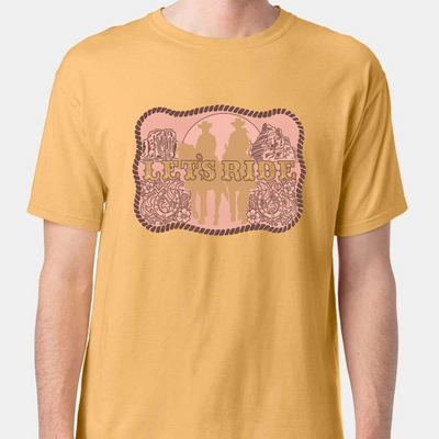 A Sunset gold colored tee with a graphic of a belt buckle. Belt buckle is a pink color with a dark brown rope border and brown western filigree on the sides with two cowboys on horses in light tan in the middle of the belt buckle and the words Let's Ride in silver across the width of the belt buckle.