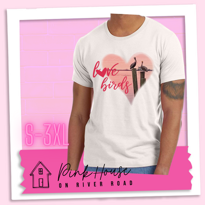 Dust tee with graphic. Graphic is a pink heart with a photo of some pelicans standing on post in the water. To the left of the heart are the words love birds in a pink cursive font and the O in love has been replaced with a heart.