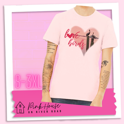 Soft Pink tee with graphic. Graphic is a pink heart with a photo of some pelicans standing on post in the water. To the left of the heart are the words love birds in a pink cursive font and the O in love has been replaced with a heart.