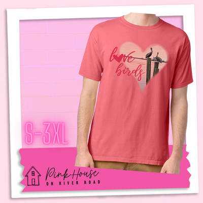 Coral tee with graphic. Graphic is a pink heart with a photo of some pelicans standing on post in the water. To the left of the heart are the words love birds in a pink cursive font and the O in love has been replaced with a heart.
