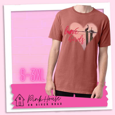 Nantucket Red tee with graphic. Graphic is a pink heart with a photo of some pelicans standing on post in the water. To the left of the heart are the words love birds in a pink cursive font and the O in love has been replaced with a heart.