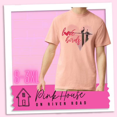 Peach tee with graphic. Graphic is a pink heart with a photo of some pelicans standing on post in the water. To the left of the heart are the words love birds in a pink cursive font and the O in love has been replaced with a heart.