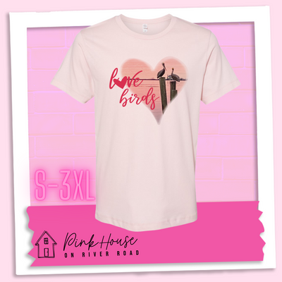 Faded Pink tee with graphic. Graphic is a pink heart with a photo of some pelicans standing on post in the water. To the left of the heart are the words love birds in a pink cursive font and the O in love has been replaced with a heart.