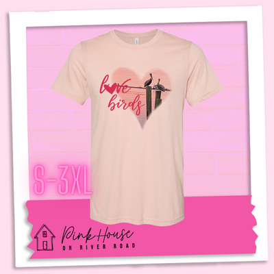 Heather peach tee with graphic. Graphic is a pink heart with a photo of some pelicans standing on post in the water. To the left of the heart are the words love birds in a pink cursive font and the O in love has been replaced with a heart.
