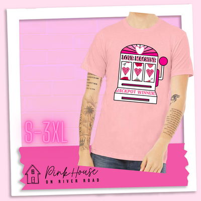 Pink Tee with a graphic. Graphic is of a white slot machine with pink accents at the top of the slot machine is an arch with different shades of pink and a heart cut out of the middle. Underneath the are are the words Love machine in hot pink with white dots to look like lights. the reels of the slot machines show three geometric hearts. The slot pull has a pink base with a white handle and pink knob on it. Under the reels are the words Jackpot Winner in pink.