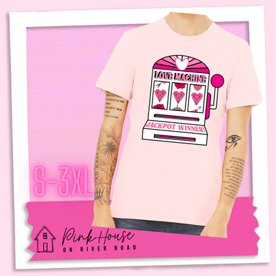 Soft Pink Tee with a graphic. Graphic is of a white slot machine with pink accents at the top of the slot machine is an arch with different shades of pink and a heart cut out of the middle. Underneath the are are the words Love machine in hot pink with white dots to look like lights. the reels of the slot machines show three geometric hearts. The slot pull has a pink base with a white handle and pink knob on it. Under the reels are the words Jackpot Winner in pink.