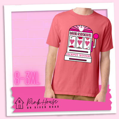 Coral Tee with a graphic. Graphic is of a white slot machine with pink accents at the top of the slot machine is an arch with different shades of pink and a heart cut out of the middle. Underneath the are are the words Love machine in hot pink with white dots to look like lights. the reels of the slot machines show three geometric hearts. The slot pull has a pink base with a white handle and pink knob on it. Under the reels are the words Jackpot Winner in pink.