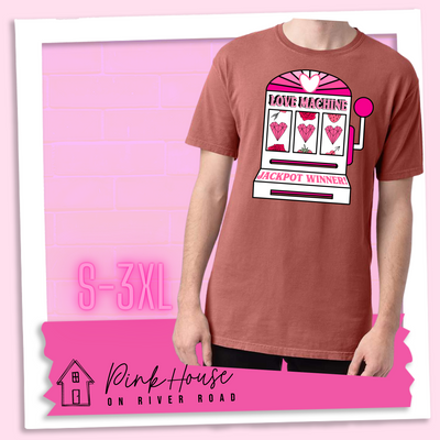 Nantucket Red Tee with a graphic. Graphic is of a white slot machine with pink accents at the top of the slot machine is an arch with different shades of pink and a heart cut out of the middle. Underneath the are are the words Love machine in hot pink with white dots to look like lights. the reels of the slot machines show three geometric hearts. The slot pull has a pink base with a white handle and pink knob on it. Under the reels are the words Jackpot Winner in pink.
