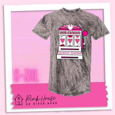 Charcoal Mineral Wash Tee with a graphic. Graphic is of a white slot machine with pink accents at the top of the slot machine is an arch with different shades of pink and a heart cut out of the middle. Underneath the are are the words Love machine in hot pink with white dots to look like lights. the reels of the slot machines show three geometric hearts. The slot pull has a pink base with a white handle and pink knob on it. Under the reels are the words Jackpot Winner in pink.