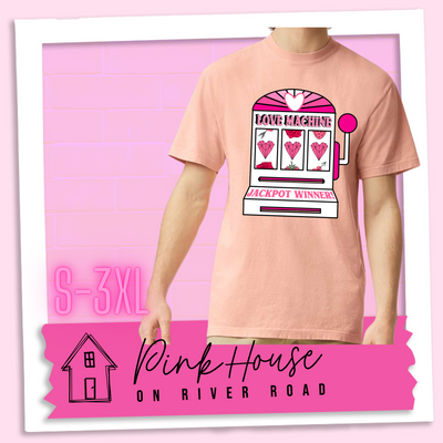 Peachy Tee with a graphic. Graphic is of a white slot machine with pink accents at the top of the slot machine is an arch with different shades of pink and a heart cut out of the middle. Underneath the are are the words Love machine in hot pink with white dots to look like lights. the reels of the slot machines show three geometric hearts. The slot pull has a pink base with a white handle and pink knob on it. Under the reels are the words Jackpot Winner in pink.