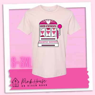 Faded Pink Tee with a graphic. Graphic is of a white slot machine with pink accents at the top of the slot machine is an arch with different shades of pink and a heart cut out of the middle. Underneath the are are the words Love machine in hot pink with white dots to look like lights. the reels of the slot machines show three geometric hearts. The slot pull has a pink base with a white handle and pink knob on it. Under the reels are the words Jackpot Winner in pink.