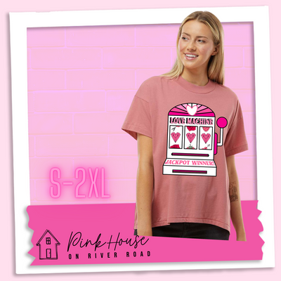 A Mauve oversized HiLo Tee with a graphic. Graphic is of a white slot machine with pink accents at the top of the slot machine is an arch with different shades of pink and a heart cut out of the middle. Underneath the are are the words Love machine in hot pink with white dots to look like lights. the reels of the slot machines show three geometric hearts. The slot pull has a pink base with a white handle and pink knob on it. Under the reels are the words Jackpot Winner in pink.