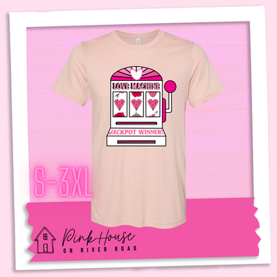 Heather Peach Tee with a graphic. Graphic is of a white slot machine with pink accents at the top of the slot machine is an arch with different shades of pink and a heart cut out of the middle. Underneath the are are the words Love machine in hot pink with white dots to look like lights. the reels of the slot machines show three geometric hearts. The slot pull has a pink base with a white handle and pink knob on it. Under the reels are the words Jackpot Winner in pink.