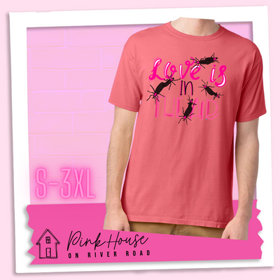 Coral Tee with Graphic. Graphic says " Love Is in the Air". "Love Is" is a hot pink cursive font with white highlights and hot pink accent lines underneath is the word "In" Is it in a black font with a hot pink highlight. on the bottom is "The Air" in Light pink with light pink accent lines. There are also black flying bugs with hot pink spots behind their heads