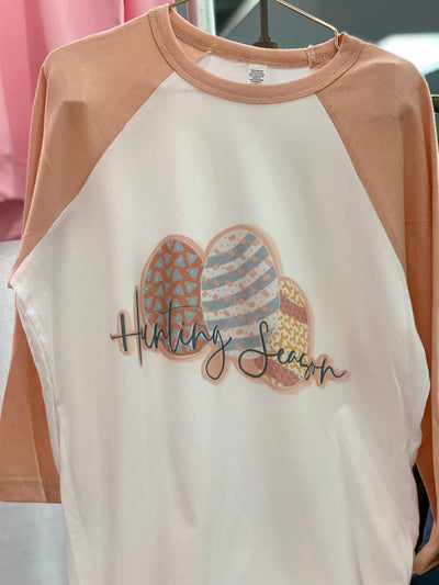 A white raglan with peach sleeves and a graphic of three differently decorated easter eggs and the words "Hunting Season" In a blue cursive font
